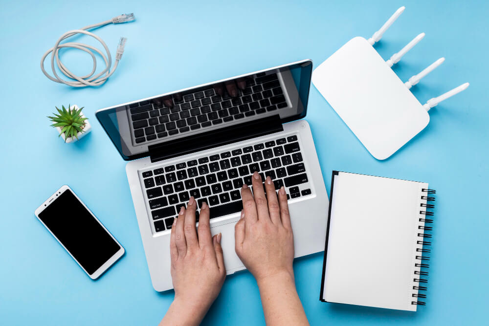 Hands typing on a laptop surrounded by a smartphone, router, notebook, ethernet cable, and small plant – representing modern digital life and assets.