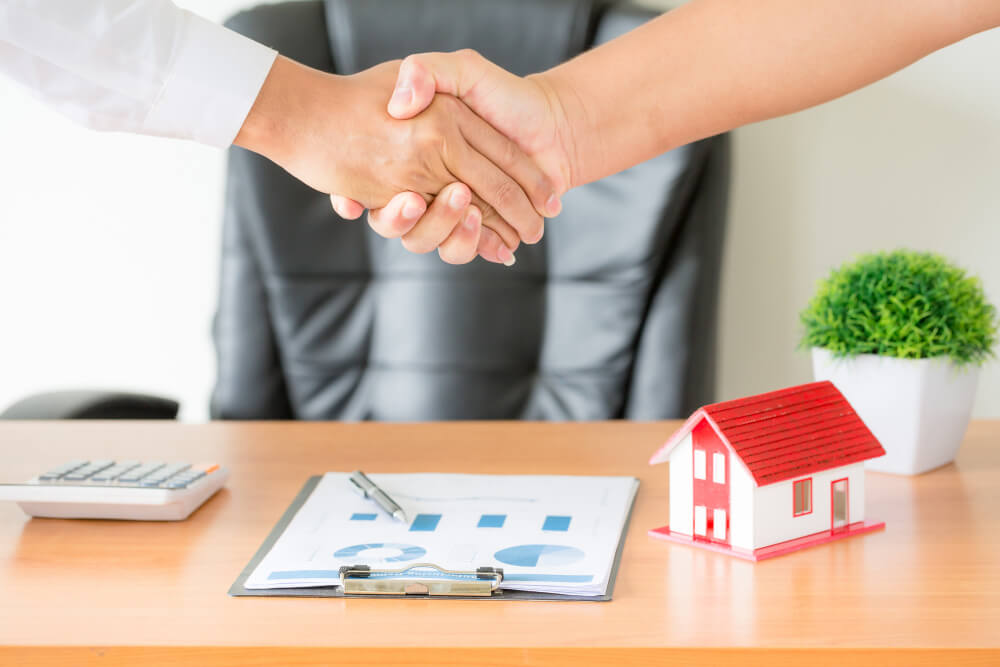 Two people shaking hands beside a house model and real estate documents, illustrating joint tenancy and tenancy in common ownership with estate planning guidance from TLR Law in Manitoba.