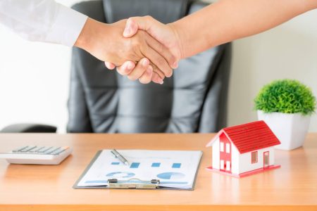 Two people shaking hands beside a house model and real estate documents, illustrating joint tenancy and tenancy in common ownership with estate planning guidance from TLR Law in Manitoba.