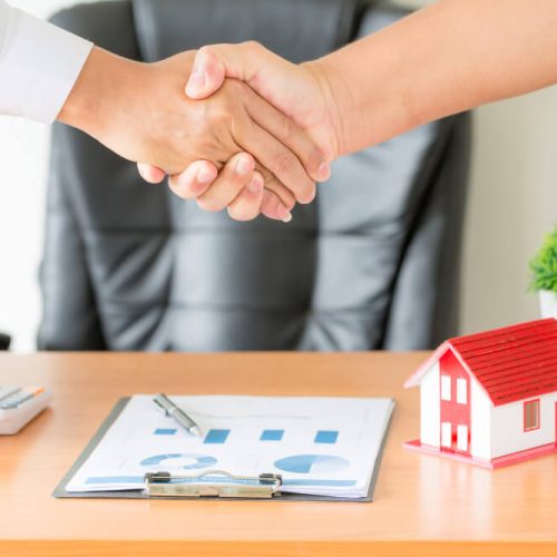 Two people shaking hands beside a house model and real estate documents, illustrating joint tenancy and tenancy in common ownership with estate planning guidance from TLR Law in Manitoba.
