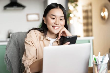 A smiling woman sits at a table in a cozy home office, looking at her smartphone while using a laptop, symbolizing digital activity. The image represents the concept of managing one’s digital legacy and online presence after death. TLR Law in Winnipeg offers guidance on what happens to your online life when you're gone.