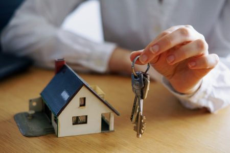 Person holding house keys beside a model home, representing the final steps of buying a house in Manitoba, including legal closing, mortgage approval, and possession with TLR Law in Winnipeg.