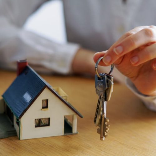 Person holding house keys beside a model home, representing the final steps of buying a house in Manitoba, including legal closing, mortgage approval, and possession with TLR Law in Winnipeg.
